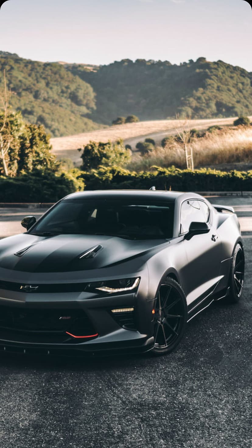 Matte black Chevrolet Camaro SS parked on an asphalt mountain road surrounded by golden grass and green hills.