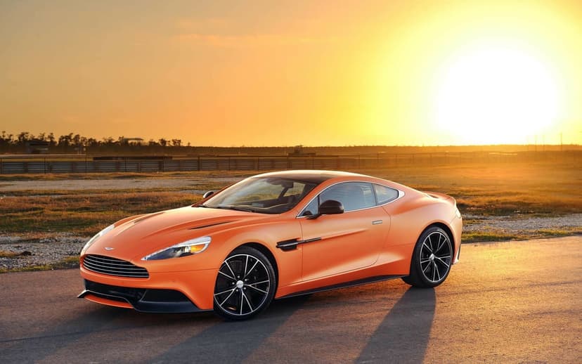 Orange luxury sports car parked on a racetrack under a bright golden sunset with long shadows on the asphalt.