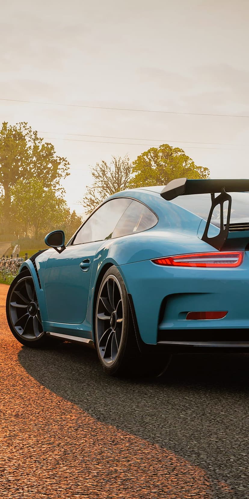 Blue Porsche 911 GT3 RS with a large rear wing parked on a gravel road during a golden sunset near green trees.