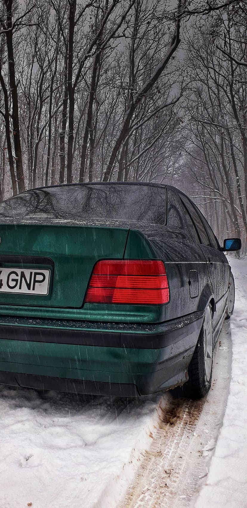 Dark green sedan parked on a snow-covered forest road with bare, frosted trees and visible tire tracks behind it.
