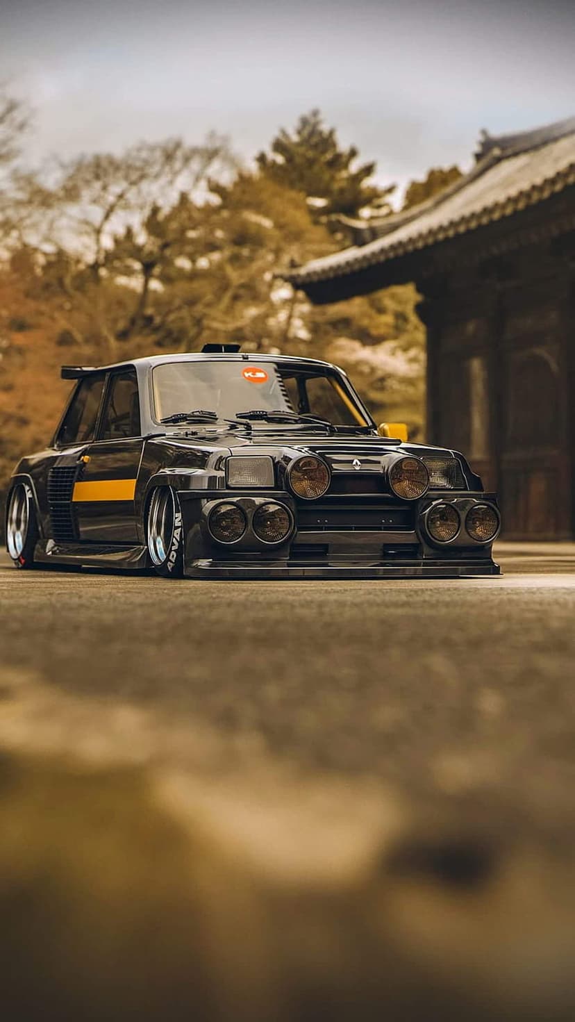 Black Renault 5 Turbo with yellow racing stripes and rally lights parked against Japanese autumn scenery.