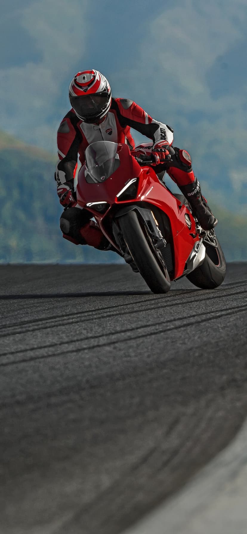 Red Ducati motorcycle leans deep into a track turn with a rider in matching red and white racing gear.