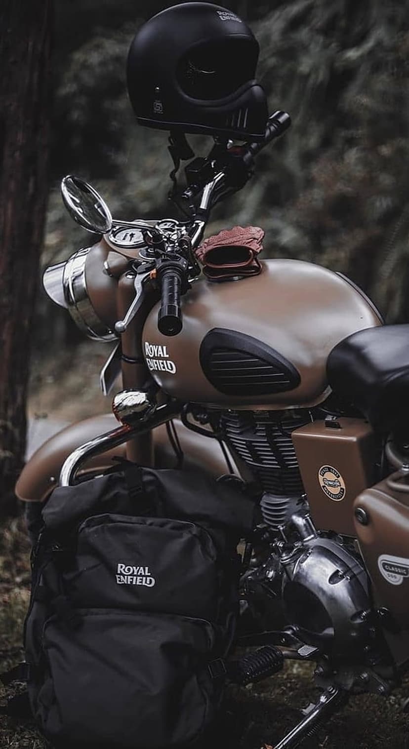 Royal Enfield motorcycle with a black helmet on the handlebars and a backpack near a lush green forest.