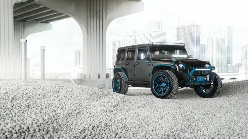Custom black Jeep Wrangler with blue accents on white gravel under a concrete bridge with a city background.