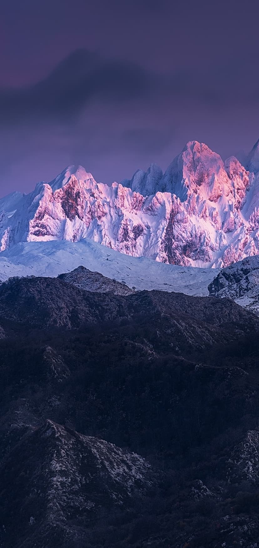 Snowy mountain peaks glowing with pink sunrise light against a purple sky above dark, shadowed valley slopes.