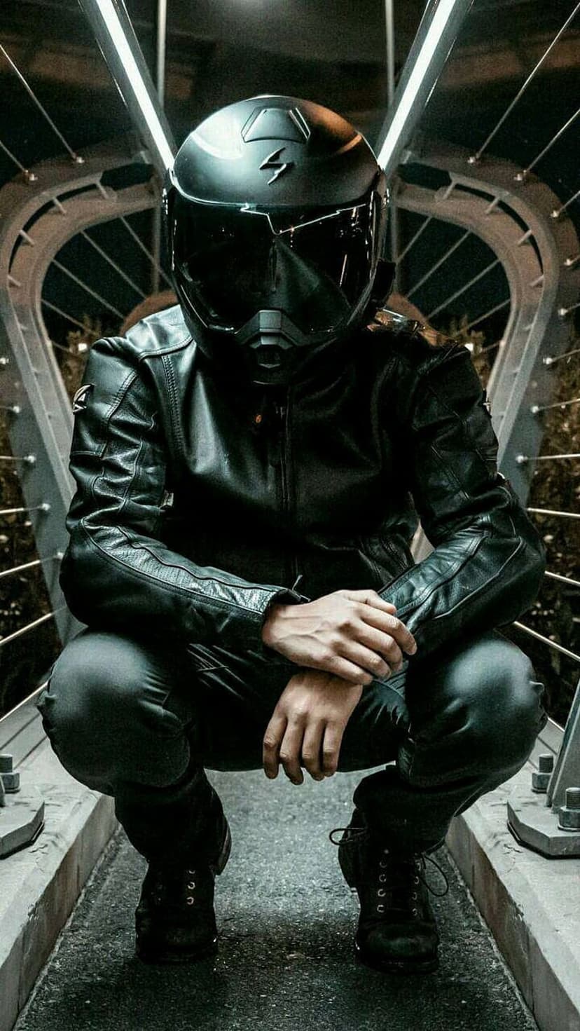 Biker in full black gear squats on an illuminated industrial bridge, creating a dark urban silhouette at night.