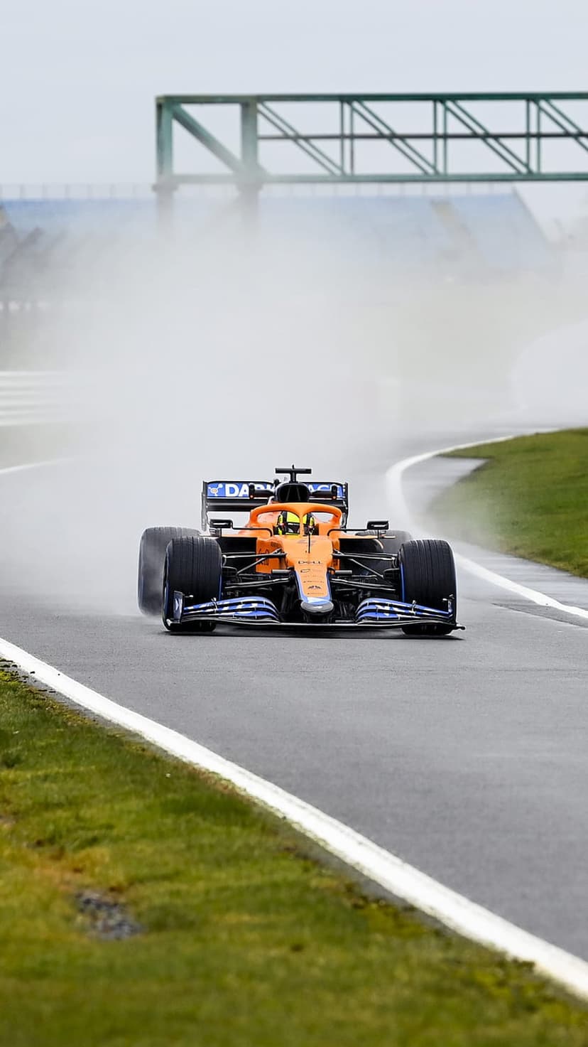 McLaren Formula One car speeds toward the viewer on a soaked track under a misty grey sky with water splashing.