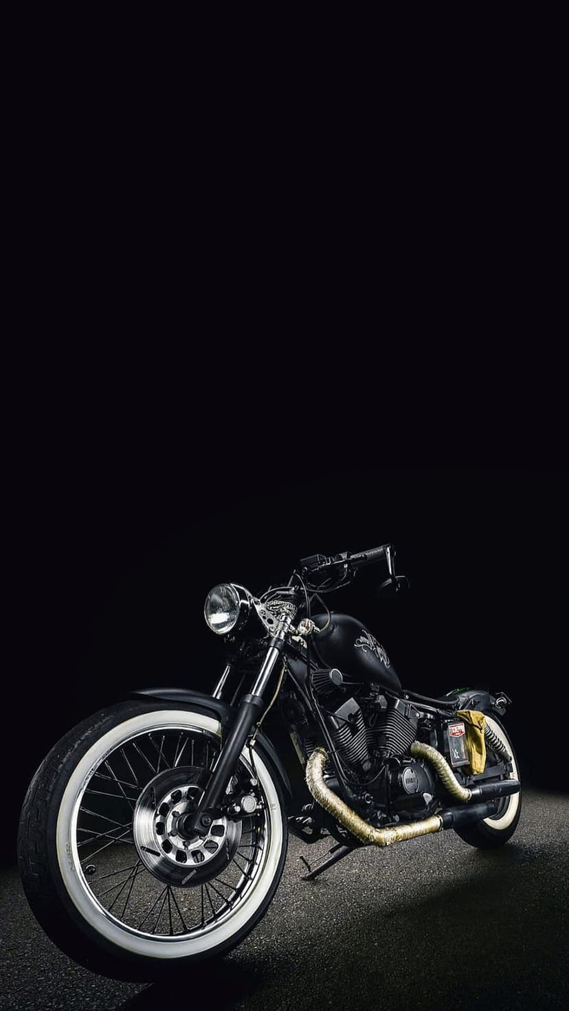 Black cruiser motorcycle featuring gold exhaust pipes and whitewall tires parked in a dark studio setting.