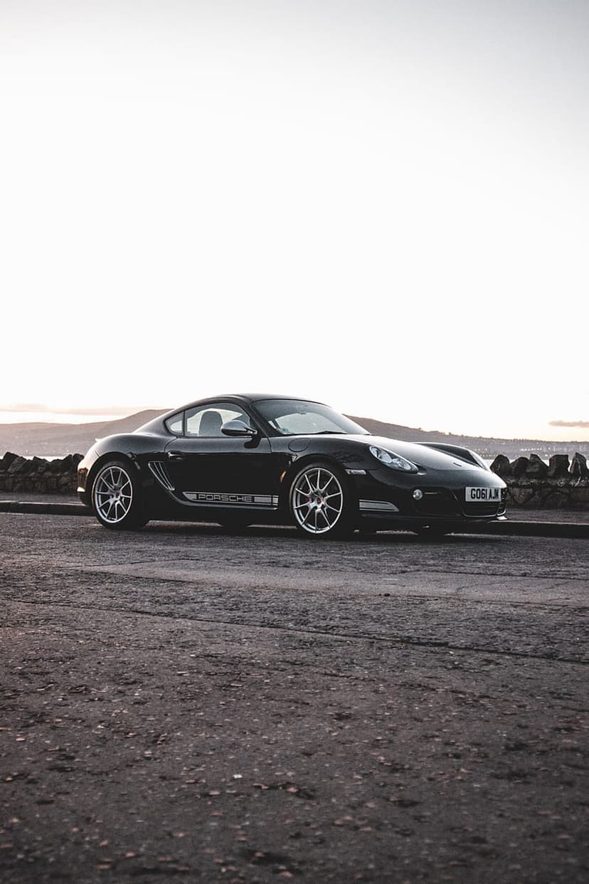 Black Porsche Cayman S parked on an asphalt road beside a stone wall and hazy coastal hills in profile view.