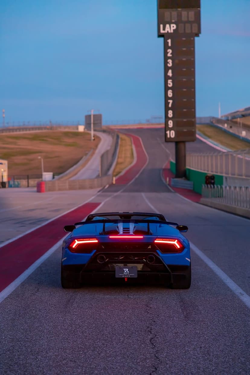 Blue Lamborghini convertible with glowing red taillights races on a track at dusk past a tall lap counter.