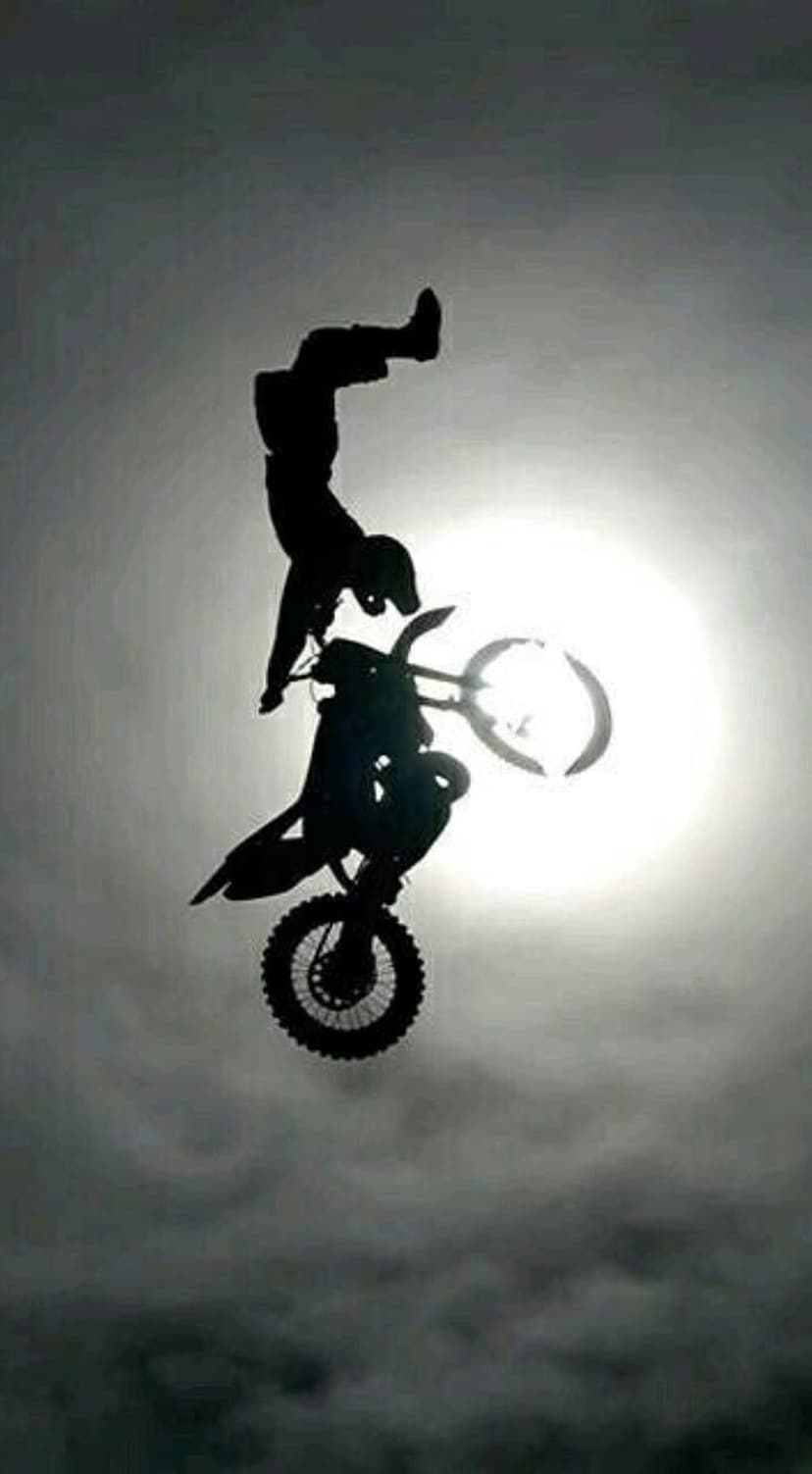 Motocross rider performing a backflip silhouette against a bright sun and dense white clouds in a blue sky.