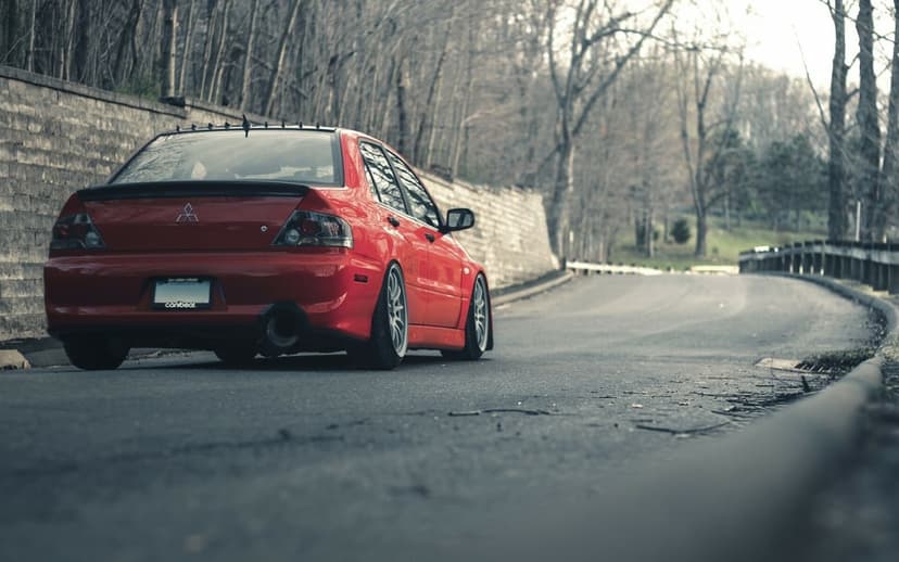Rear view of a red Mitsubishi Lancer Evolution on a gray asphalt road beside a stone wall and bare trees.