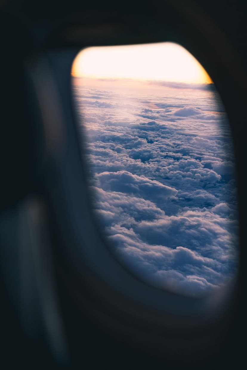 An oval airplane window frames a vibrant sea of pink and orange sunset clouds above a deep blue horizon line.