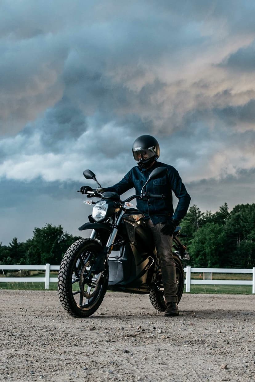 Rider on a black motorcycle under a vast sky of dark storm clouds next to a white fence in a rural field.