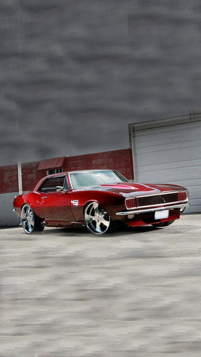 Red 1967 Chevrolet Camaro with chrome wheels parked against a brick wall in a vertical mobile orientation.