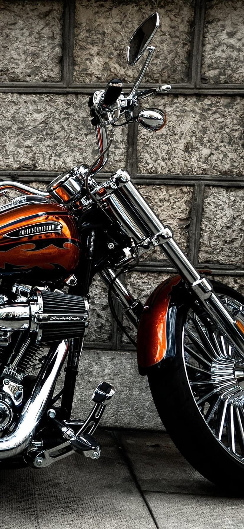 Harley-Davidson motorcycle with orange flame paint and chrome forks parked against a textured gray stone wall.