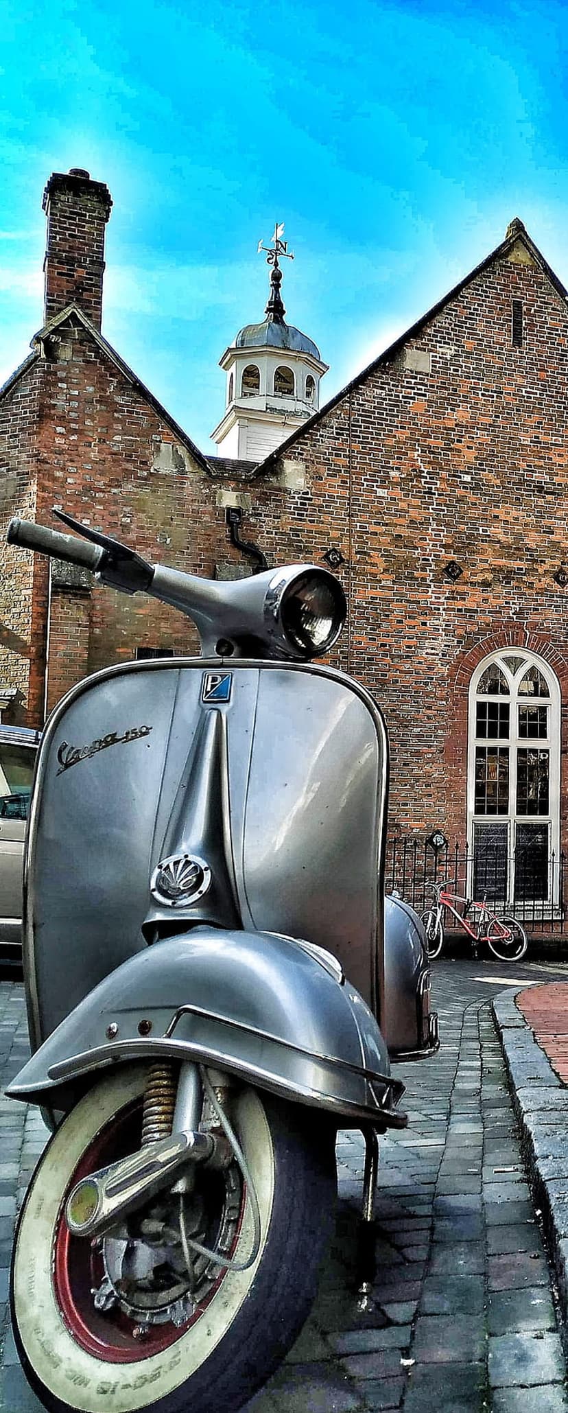 Silver Vespa scooter on cobblestones in front of a red brick tower with an arched window and clear blue sky.