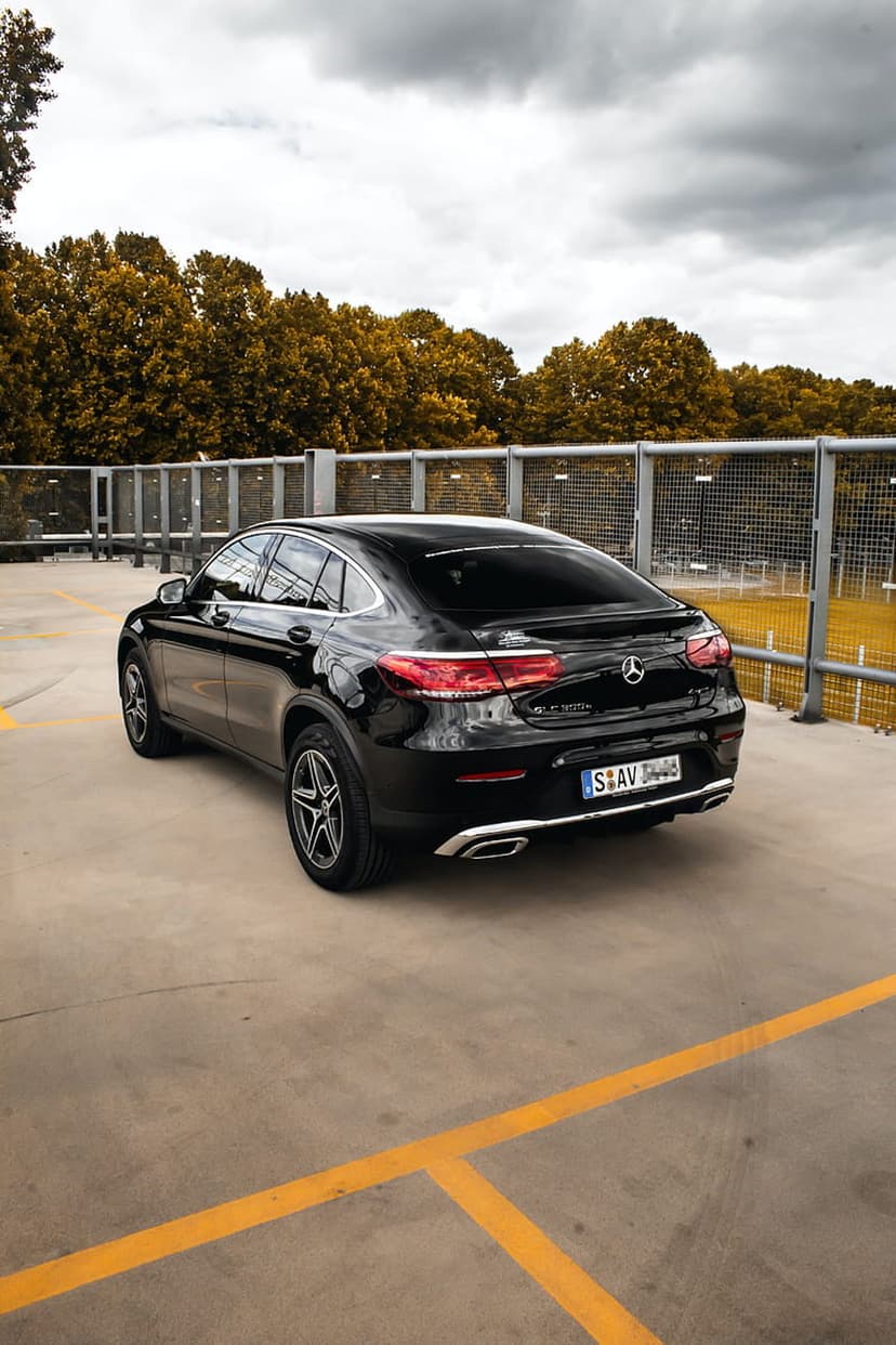 Mercedes GLC Coupe in black parked on concrete with yellow lines, reflecting autumn trees and a cloudy sky.