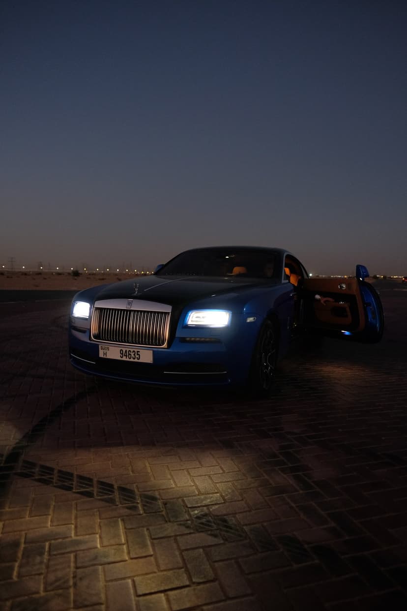 Blue Rolls-Royce coupe with an open door sits on a paved road against a twilight desert and distant city lights.