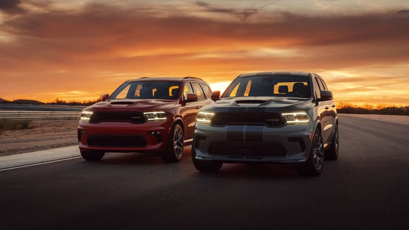 Two Dodge Durango SRT SUVs on an asphalt track under a vibrant orange and red sunset sky with glowing headlights.