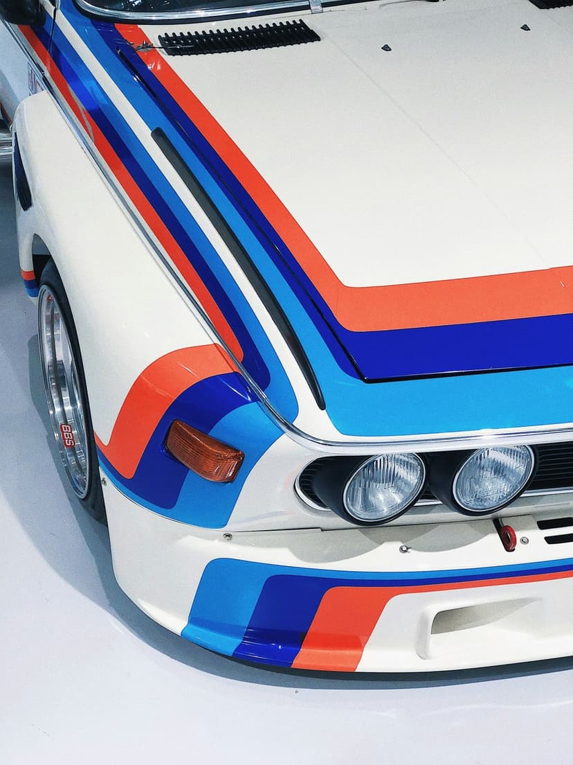 White BMW race car front featuring iconic blue and orange M stripes, round headlights, and flared fenders.