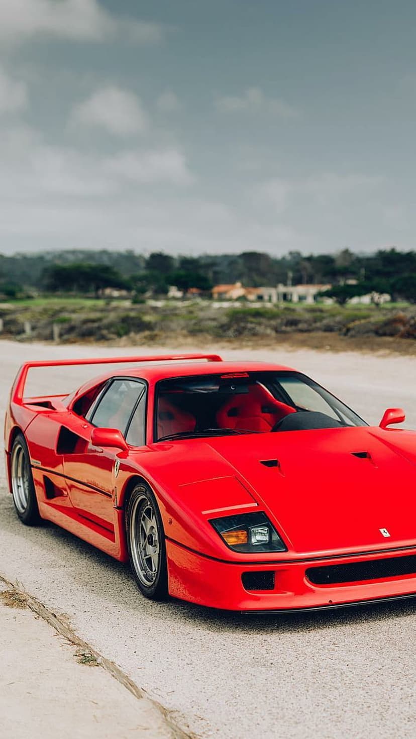 Red Ferrari F40 supercar parked on a concrete road with a tall rear wing against a cloudy coastal backdrop.
