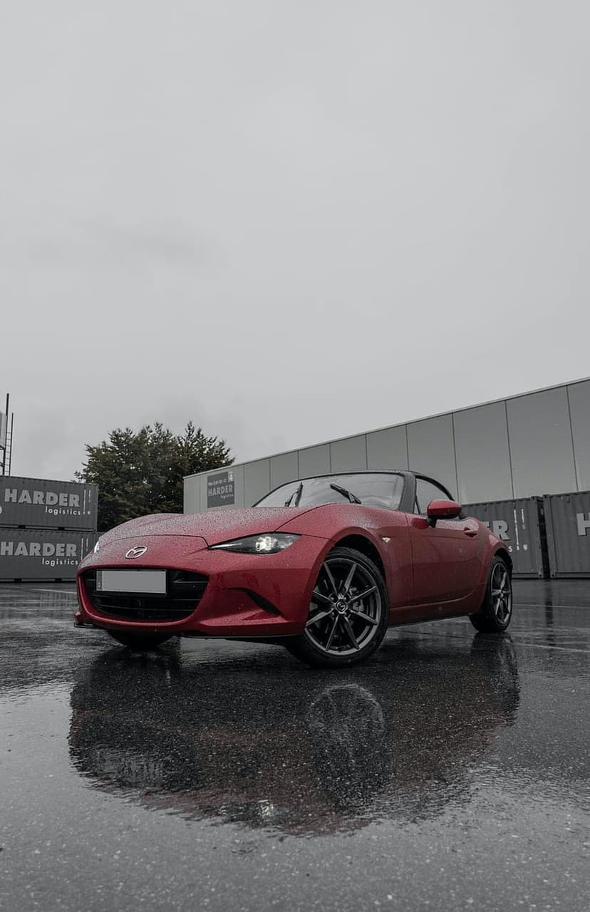 Red Mazda MX-5 convertible parked on wet asphalt with mirror reflections near industrial shipping containers.