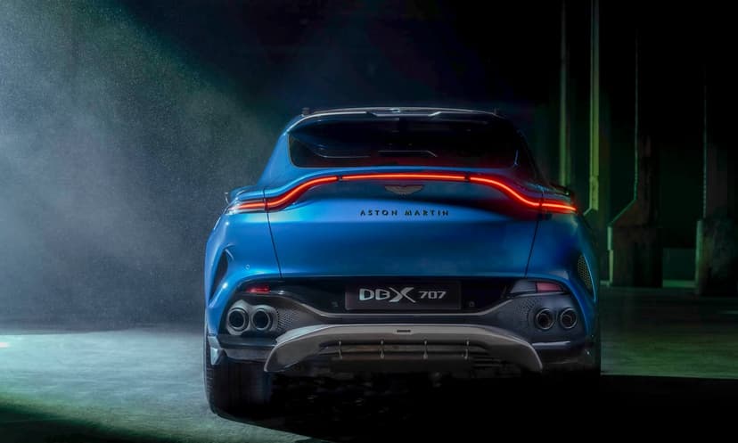 Rear view of a blue Aston Martin DBX 707 SUV with curved red LED taillights in a dark industrial space.