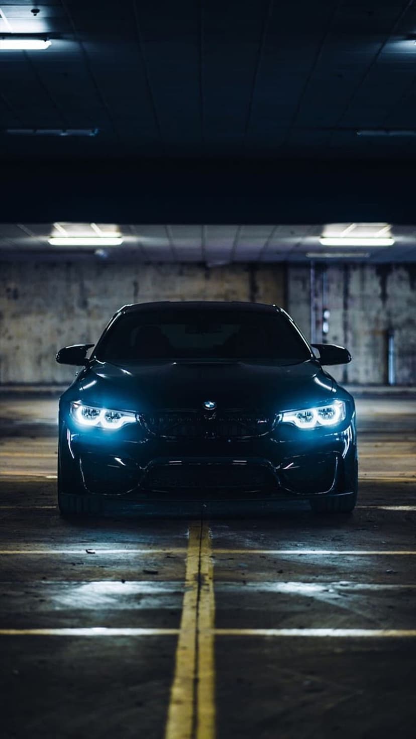 Black BMW M4 with glowing blue headlights parked on a concrete garage floor marked with yellow safety stripes.