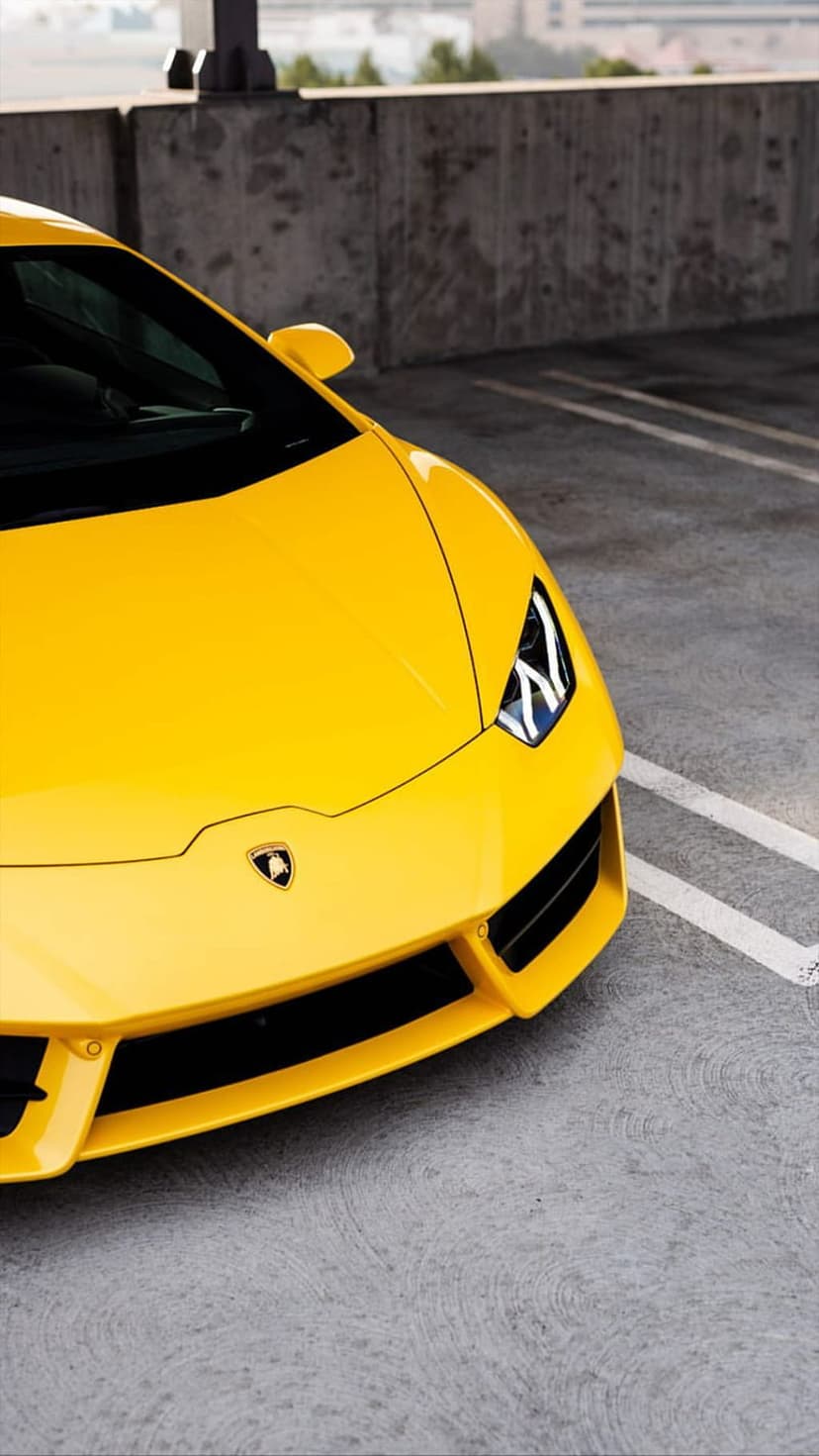 Yellow Lamborghini supercar front view on gray concrete pavement against a tall textured brutalist wall.