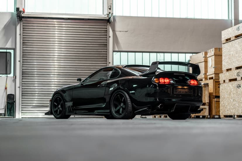 Black Toyota Supra with a large rear spoiler parked in an industrial garage next to wooden cargo crates.