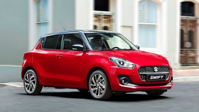 Red Suzuki Swift hatchback driving on a city street with motion blur and modern architecture background.