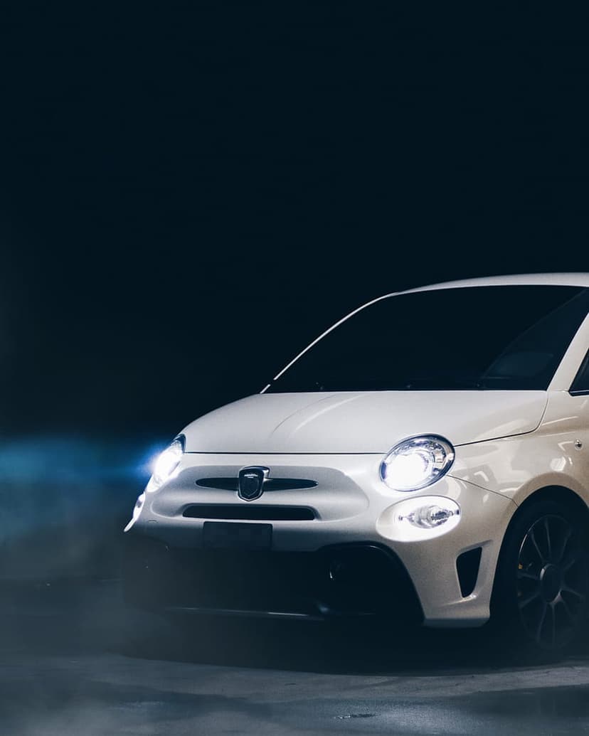 White Fiat 500 Abarth headlights glow brightly against a dark, smoky backdrop in a vertical automotive close-up.
