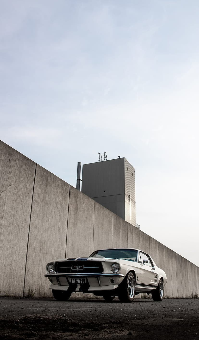 White classic Ford Mustang with blue racing stripes parked against a gray textured concrete wall in an urban city.