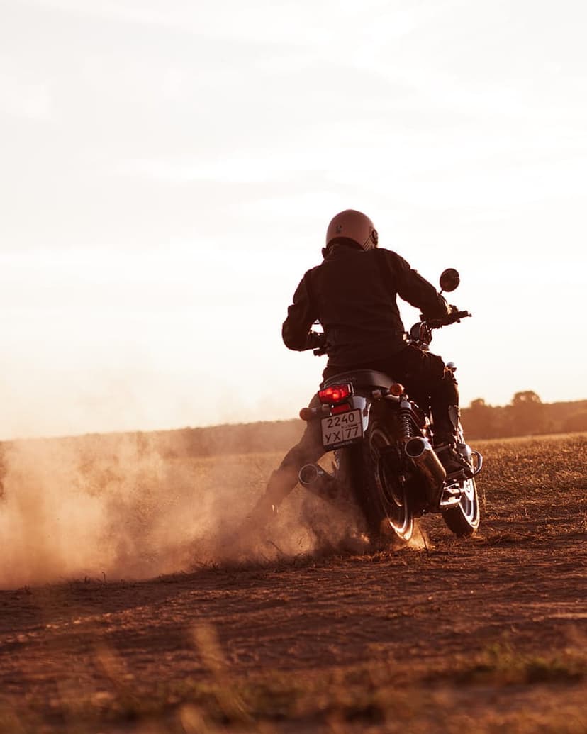 Motorcyclist in a leather jacket kicks up a massive dust cloud while riding a dirt bike during a golden sunset.
