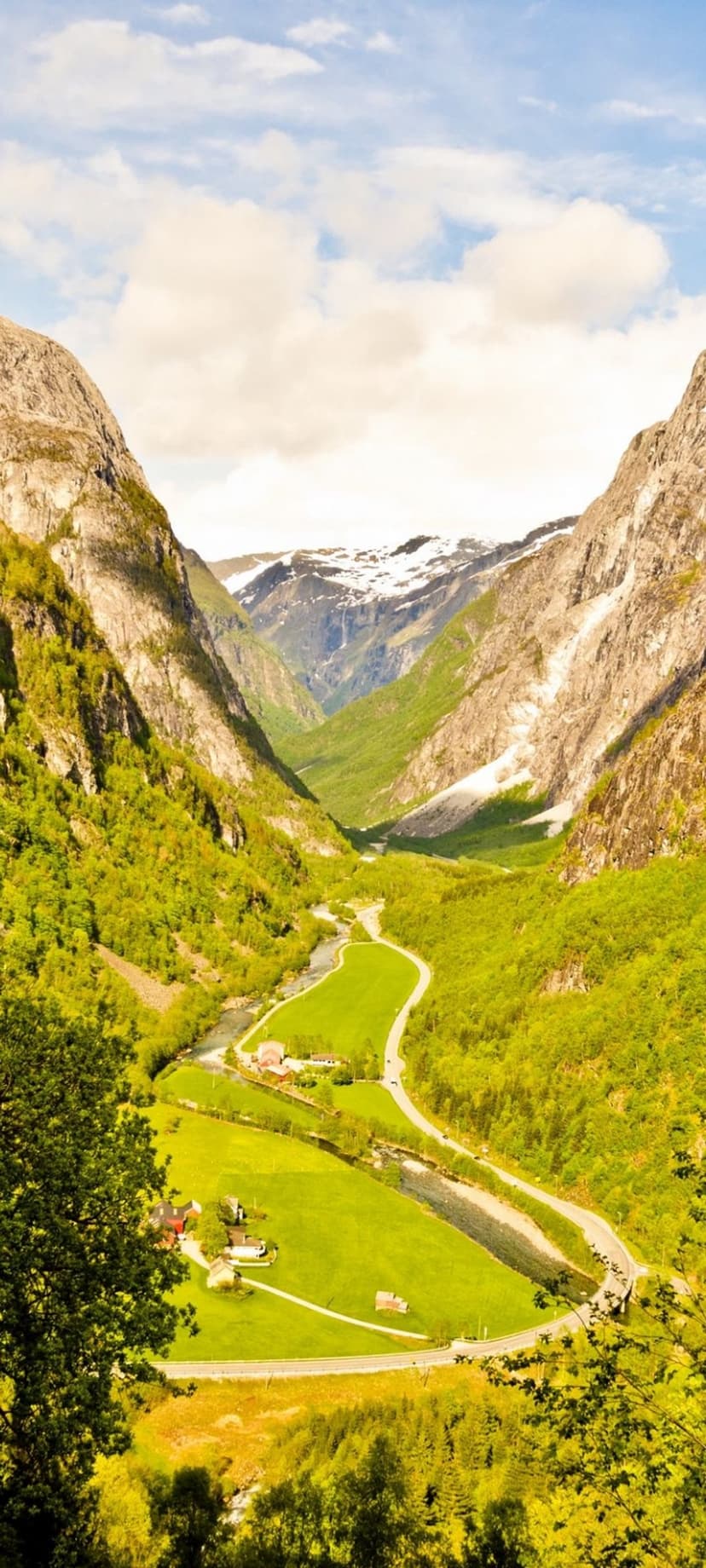 Lush green valley in Norway with a winding river nestled between steep, snow-capped mountains under a soft sky.