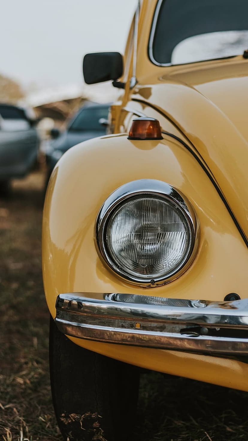 Yellow Volkswagen Beetle headlight and chrome bumper reflecting an overcast sky at a classic car show gathering.