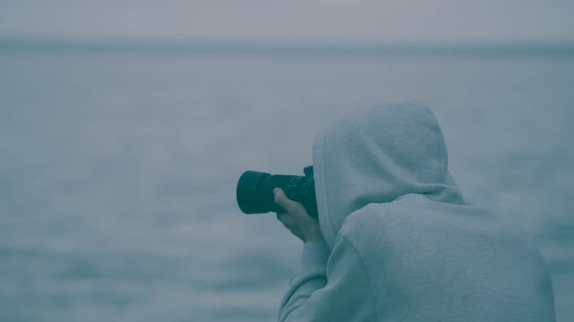 Photographer in a grey hoodie stands at the water's edge holding a camera toward a misty, overcast ocean horizon.