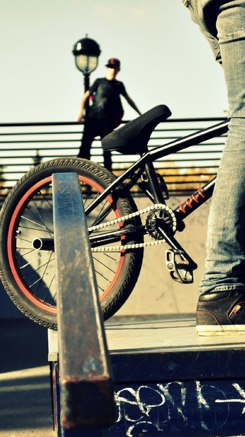 BMX rider in a cap stands beside a black bike with red rims at a skatepark under an urban street lamp.