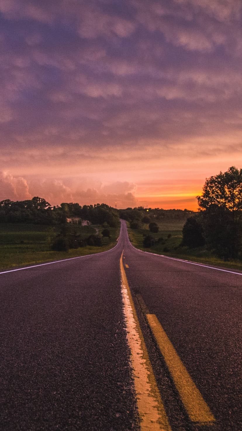 Paved highway leading toward a vibrant orange and purple sunset sky with green fields and distant trees.