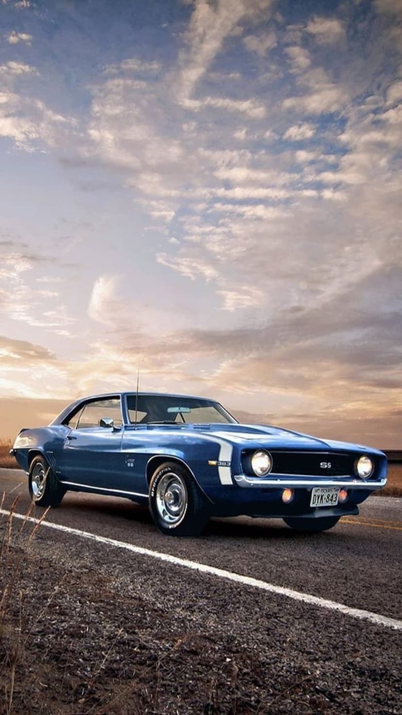 Blue Camaro SS muscle car parked on a rural road under a dramatic golden sunset with glowing orange clouds.