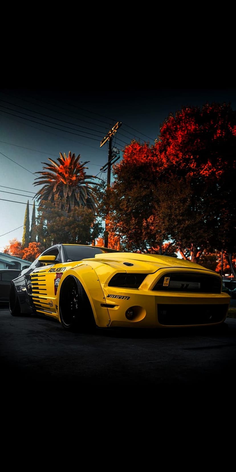 Yellow widebody Ford Mustang with racing decals parked on a suburban street lined with palm trees and red leaves.