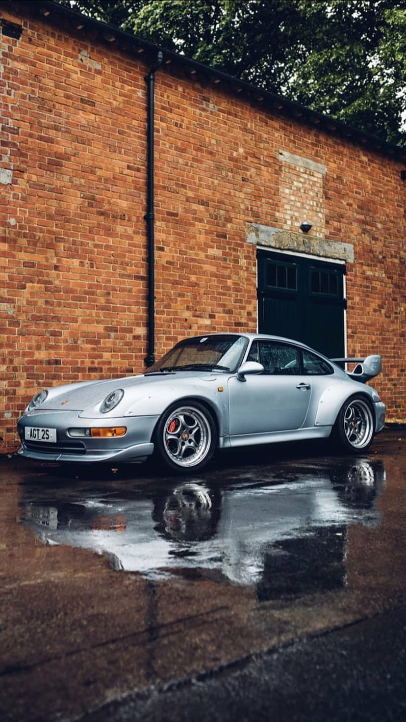Silver Porsche 911 Turbo 993 with a rear spoiler reflects on wet asphalt in front of a rustic red brick wall.