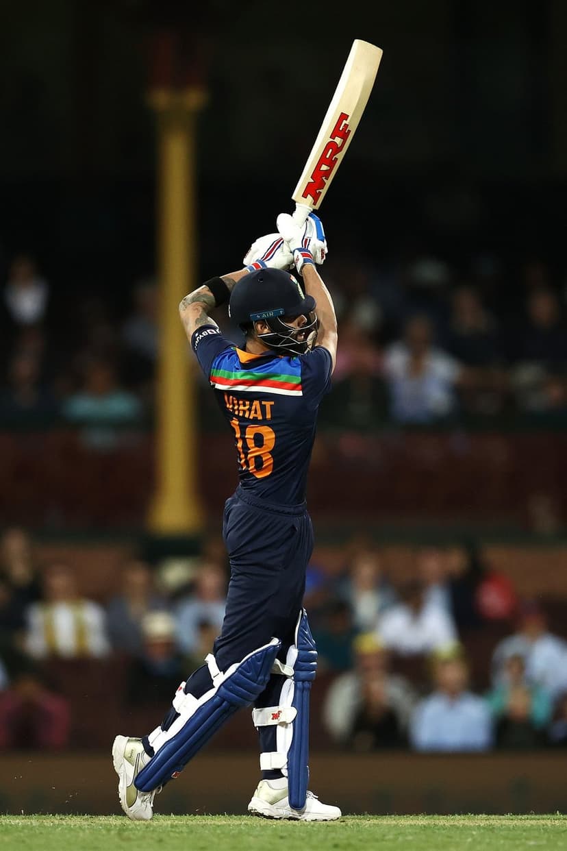 Virat Kohli wearing a blue cricket uniform swings his bat against a blurred stadium crowd during a live match.