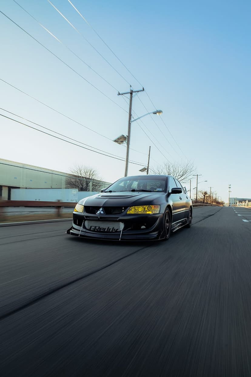 Black Mitsubishi Lancer Evolution X with yellow headlights and intercooler driving on a sunny paved road.