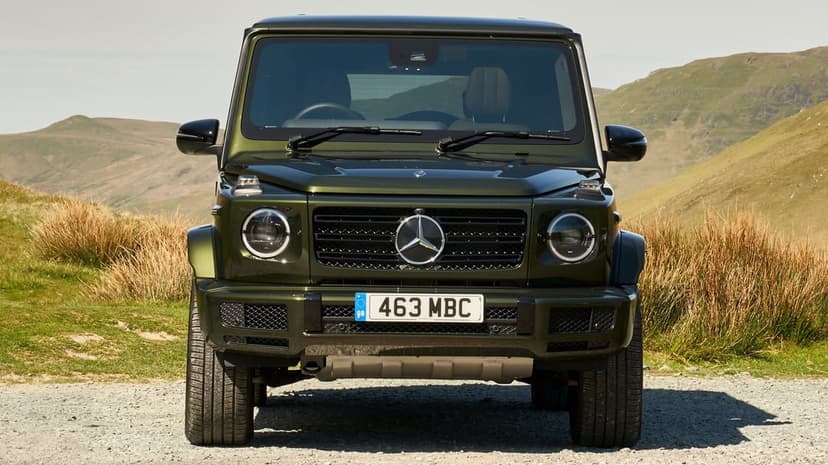 Front view of a dark green Mercedes-Benz G-Class parked on a gravel road with rolling hills in the background.