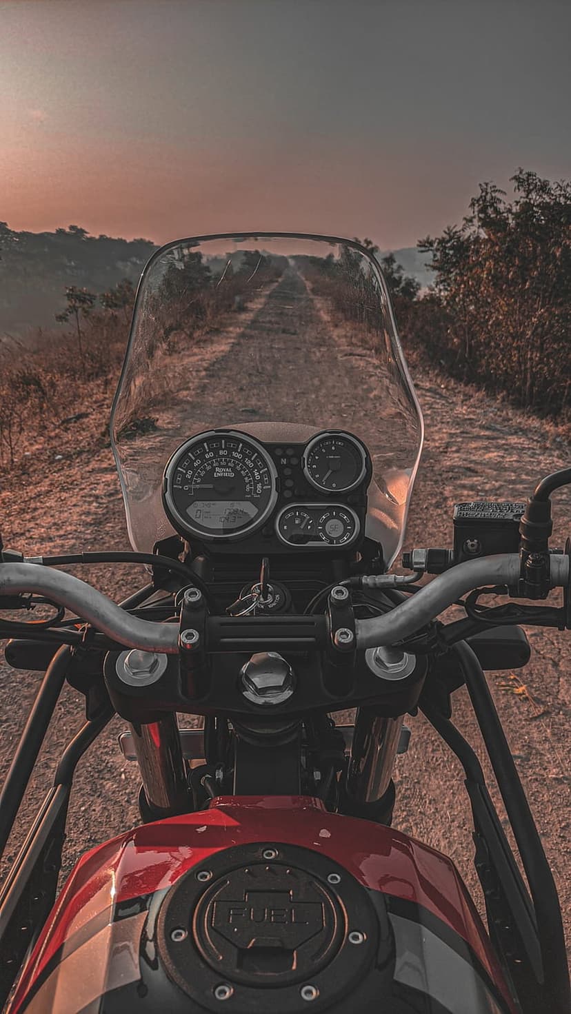 Motorcycle dashboard and fuel tank face a dusty dirt road under a glowing golden hour sky in a POV shot.