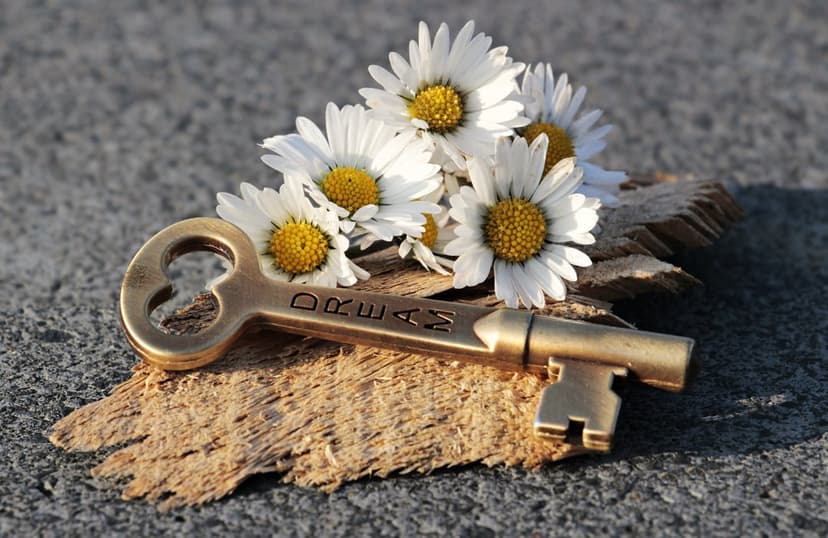 Golden key with engraved dream text resting on wood next to white daisies against a grey textured surface.
