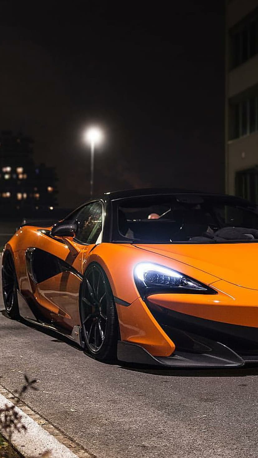 Orange McLaren supercar with lit headlights parked on a dark asphalt road amidst an urban nighttime setting.