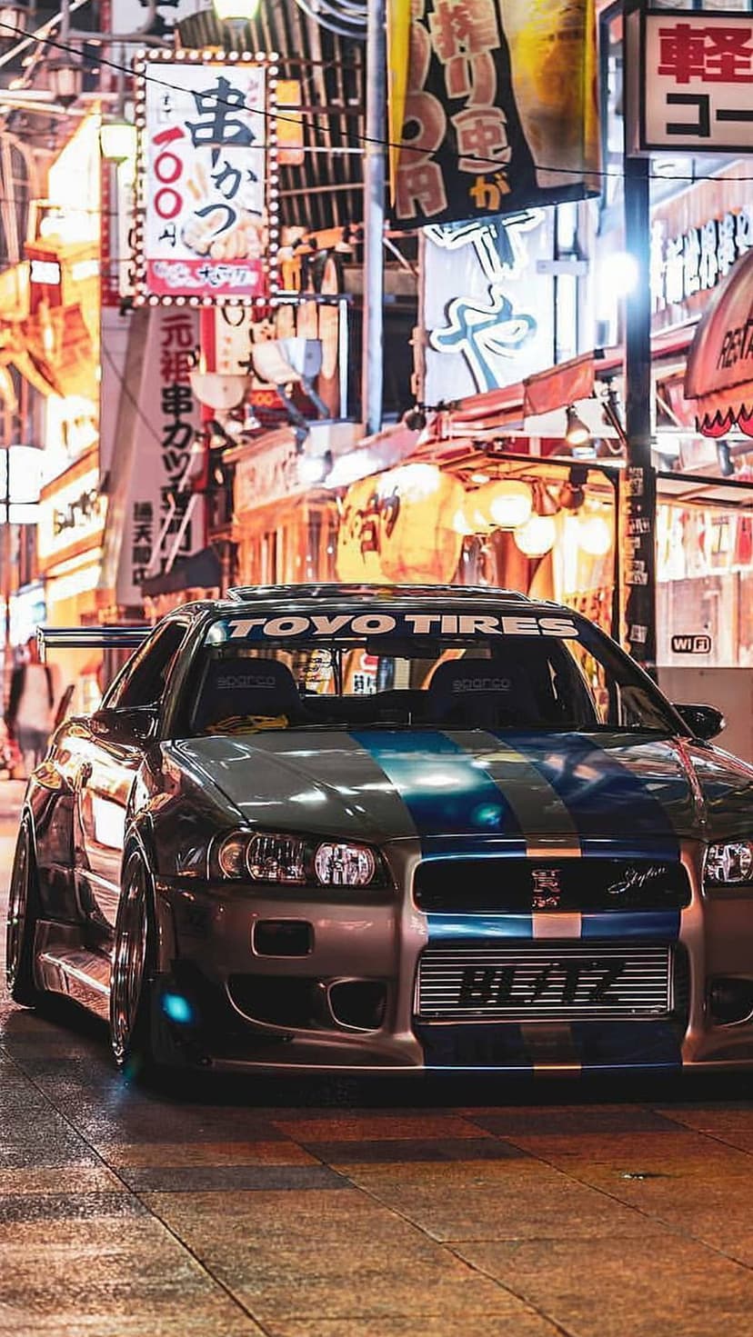 Customized sports car with racing tires parked on a wet cobblestone street under glowing Japanese neon signs.